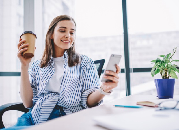 A smiling young woman, sitting at a desk, holding a cup of coffee, while looking at her mobile device.